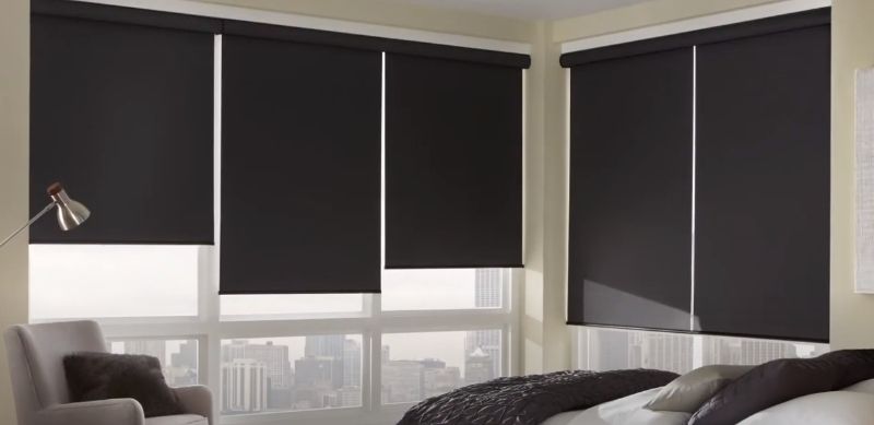 A high-rise bedroom with brown roller shades looks out over a city view. 