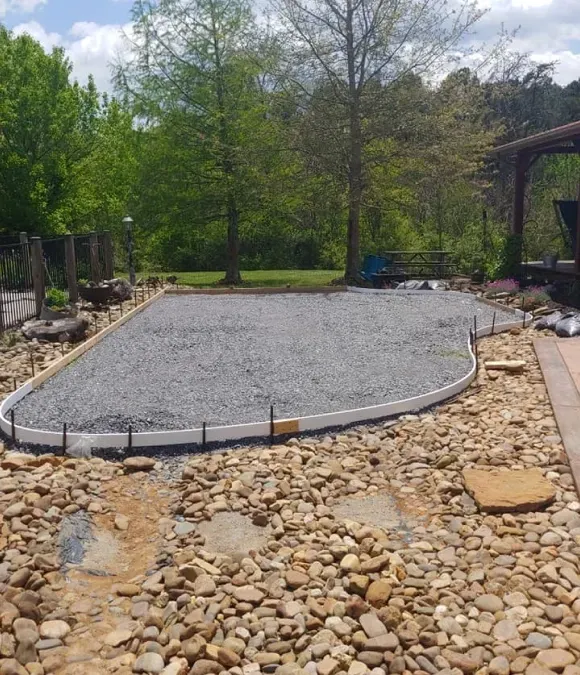 Gravel patio under construction beside a stone border and trees in a backyard.