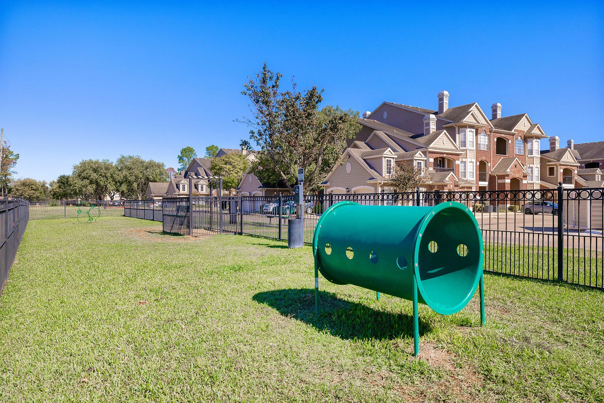 Green cylindrical play tunnel in a fenced grassy area with apartment buildings in the background.