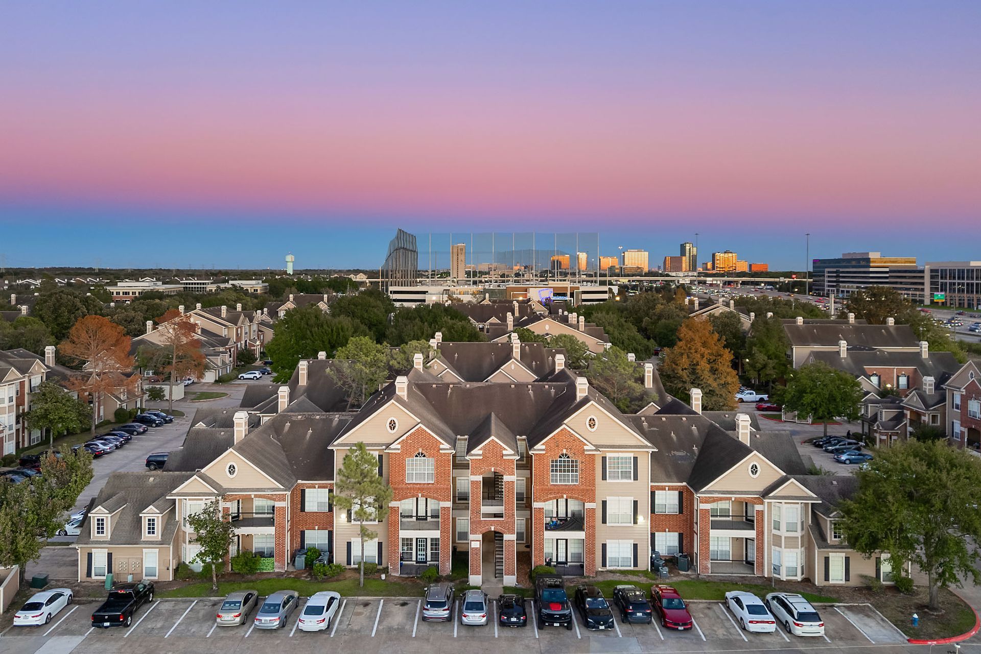 Aerial view of a multi-building apartment complex with a parking lot and city skyline in the distance.