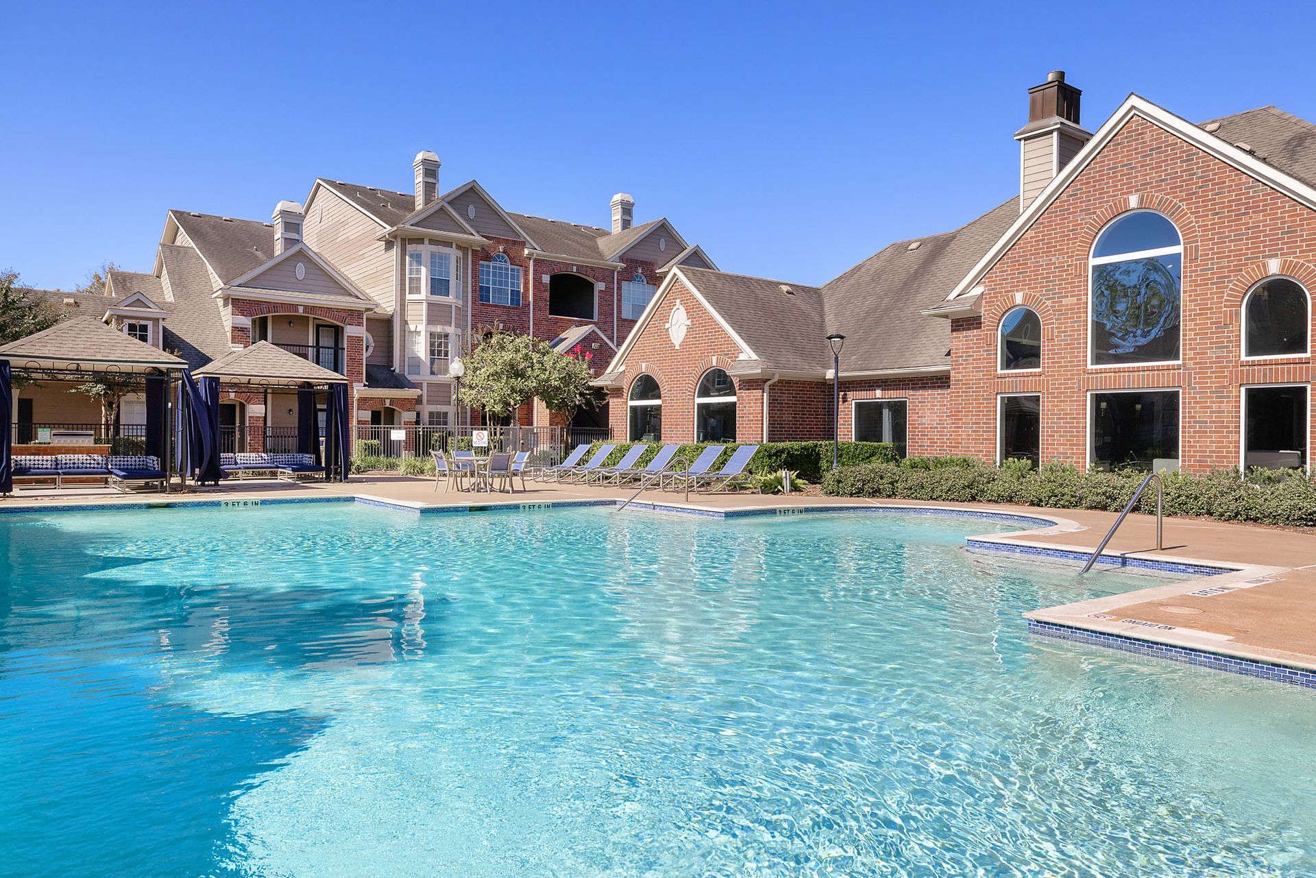 Outdoor pool area of a multifamily community with lounge chairs and brick buildings.