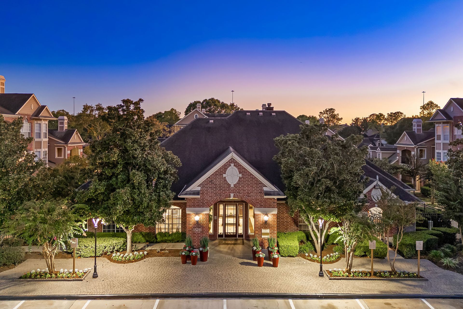 Brick clubhouse entrance with arched doors, lamps, and greenery at dusk