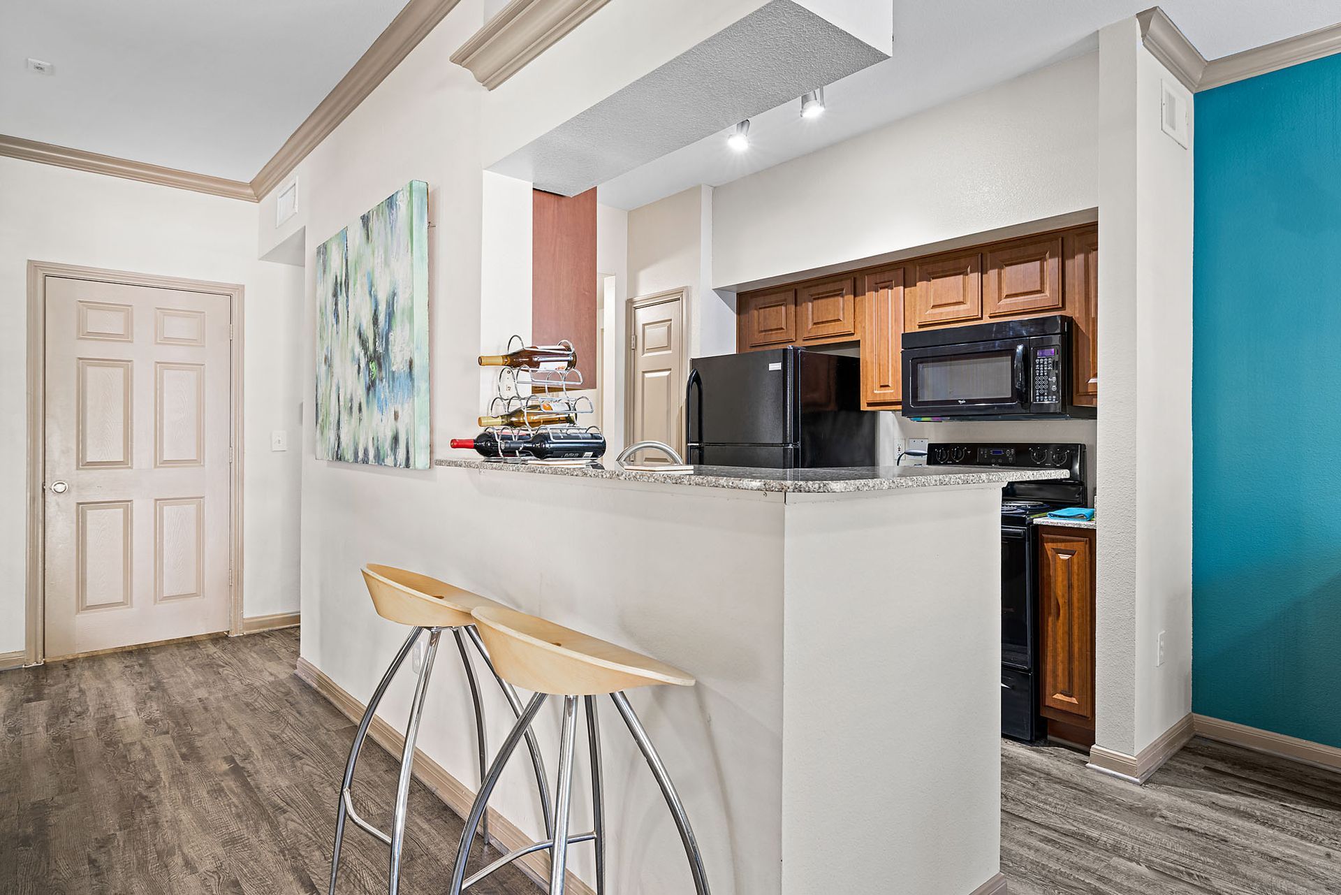 Kitchen in an apartment with a granite bar counter, two stools, stainless appliances, and teal accent wall.