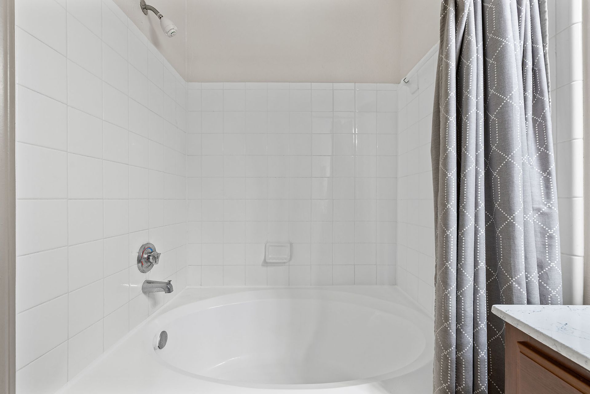 White-tiled bathroom with bathtub, chrome faucet, and grey patterned shower curtain.