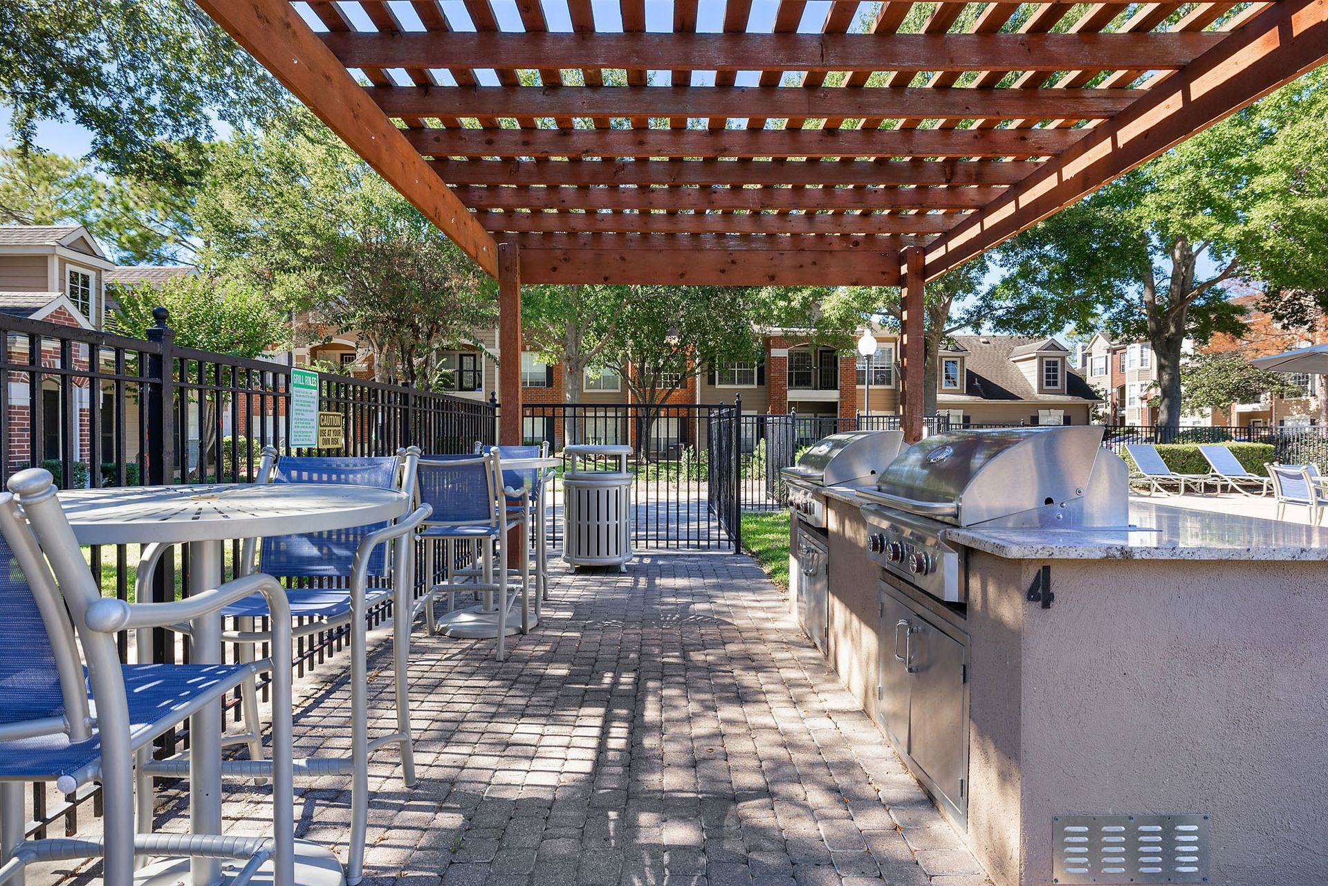Outdoor communal grilling area under a wooden pergola with blue chairs and stainless grills.