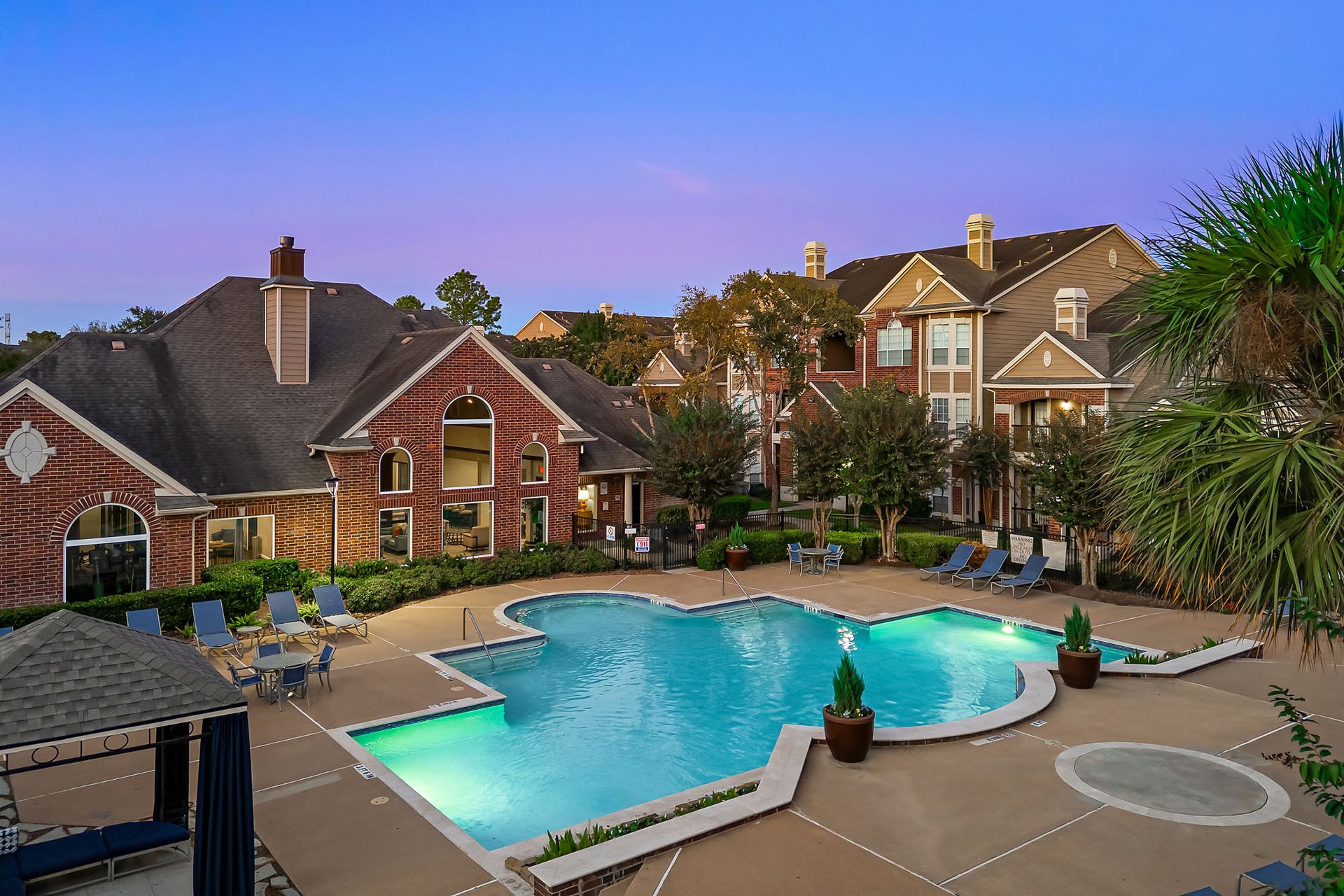 Outdoor pool area at an apartment community with lounge chairs and brick residential buildings in the background.