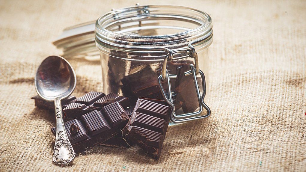 A jar filled with chocolate next to a bar of chocolate and a spoon.