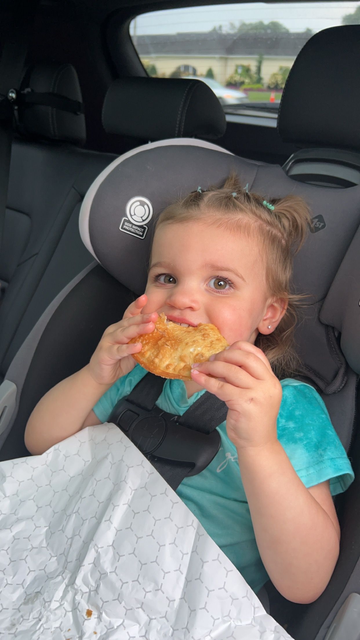 A little girl is sitting in a car seat eating a donut.