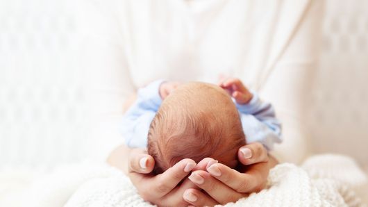 Hände umfassen den Hinterkopf eines Neugeborenen, das Baby trägt blau-weiße Kleidung, weichgezeichnet.