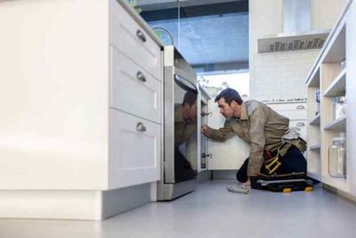 A repairman kneeling in a white kitchen, inspecting an oven. He wears a work belt and has his tools nearby.