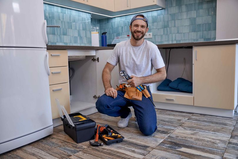 A smiling plumber kneels in a kitchen, holding a wrench. He wears a tool belt, a white shirt. A toolbox is open on the floor.