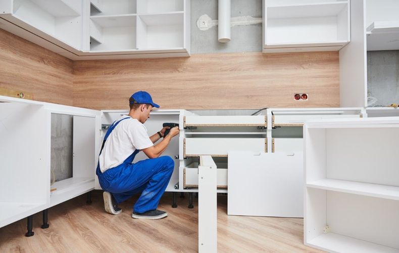 Worker kneeling in a white kitchen, measuring a cabinet with a tape measure.