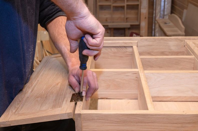 Hands using a chisel to carve a wooden cabinet door panel in a workshop