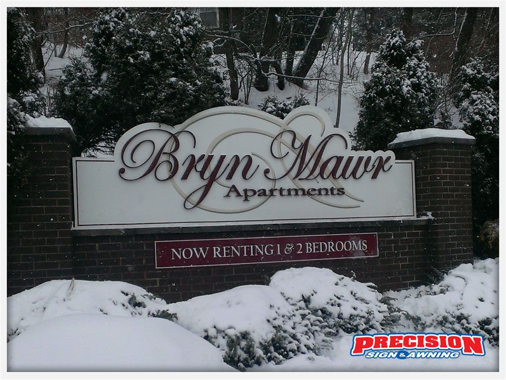 Sign for Bryn Mawr Apartments in snow; red brick pillars, white sign with maroon lettering, winter scene.