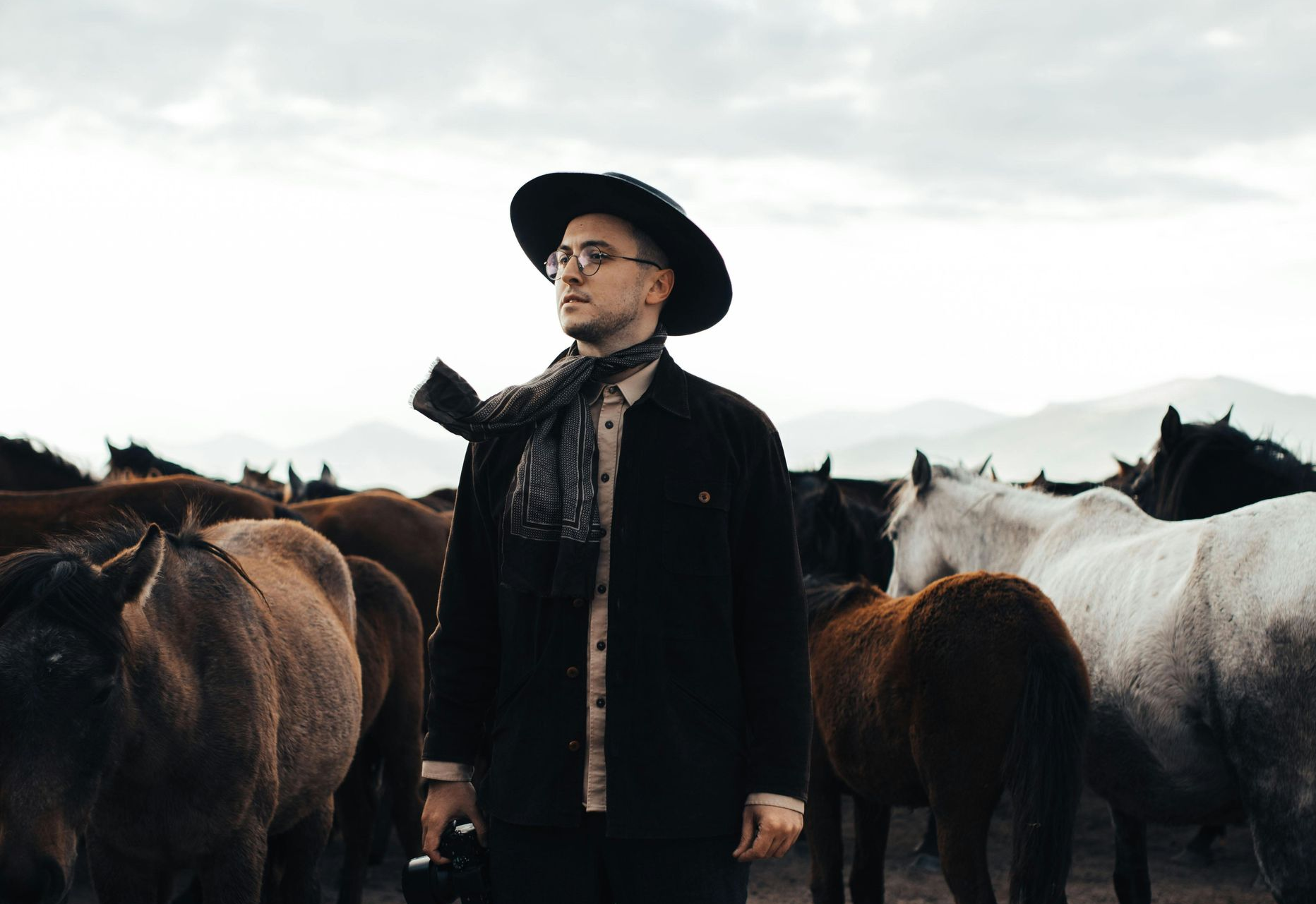 Man in a hat and scarf stands among horses on a dusty plain under a cloudy sky.