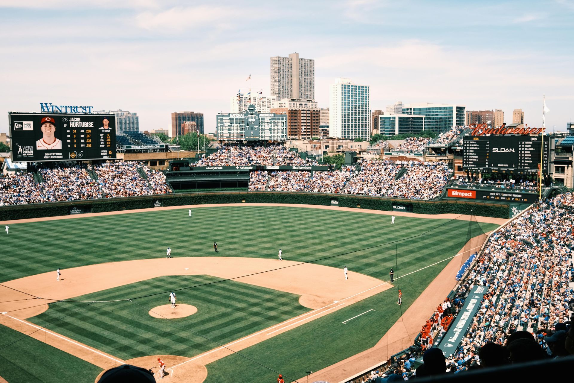 Baseball stadium filled with spectators; players on the field. Green grass, blue sky, city skyline in background.