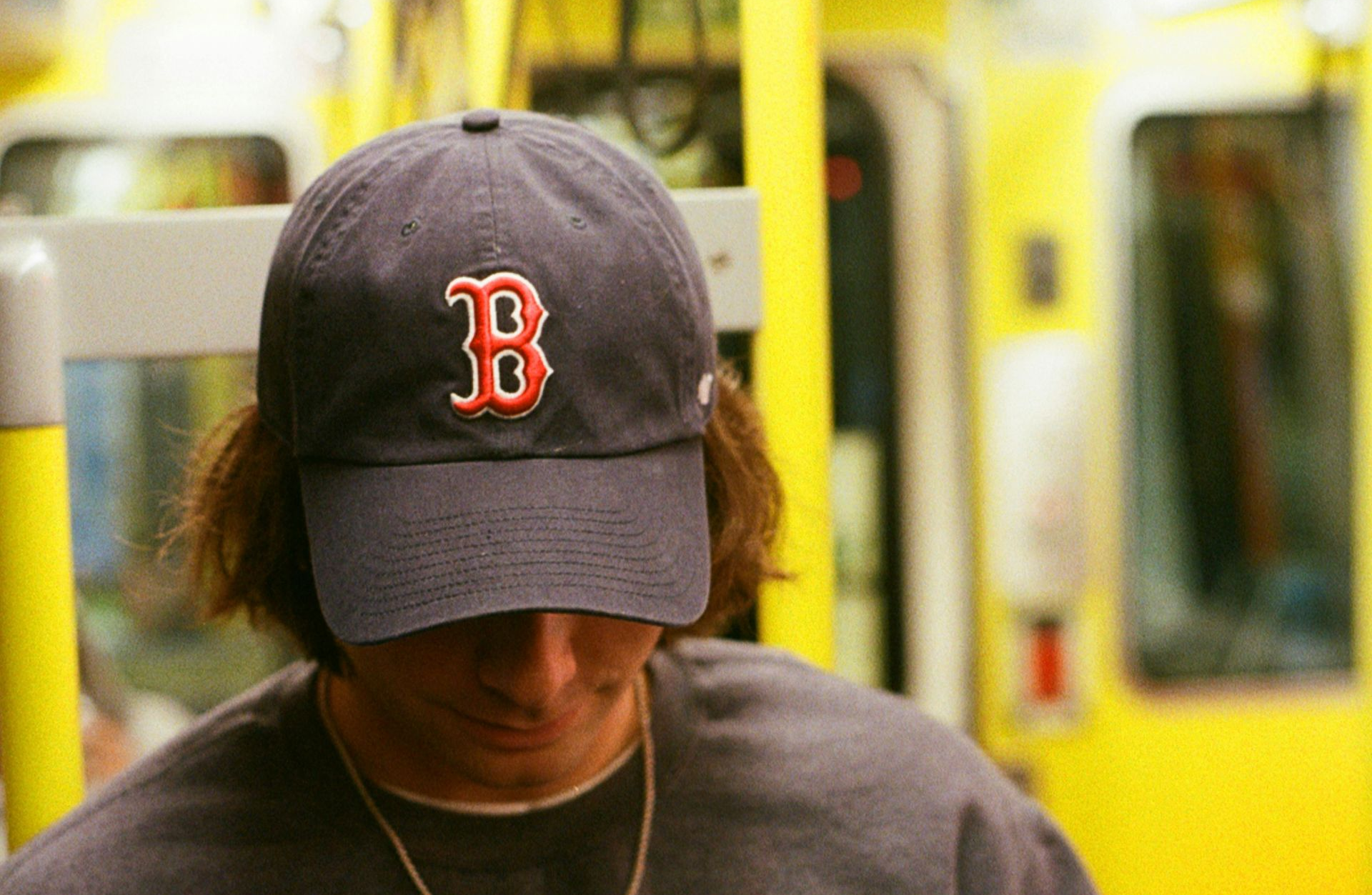 Person wearing a Boston Red Sox cap on a yellow subway train, looking down.