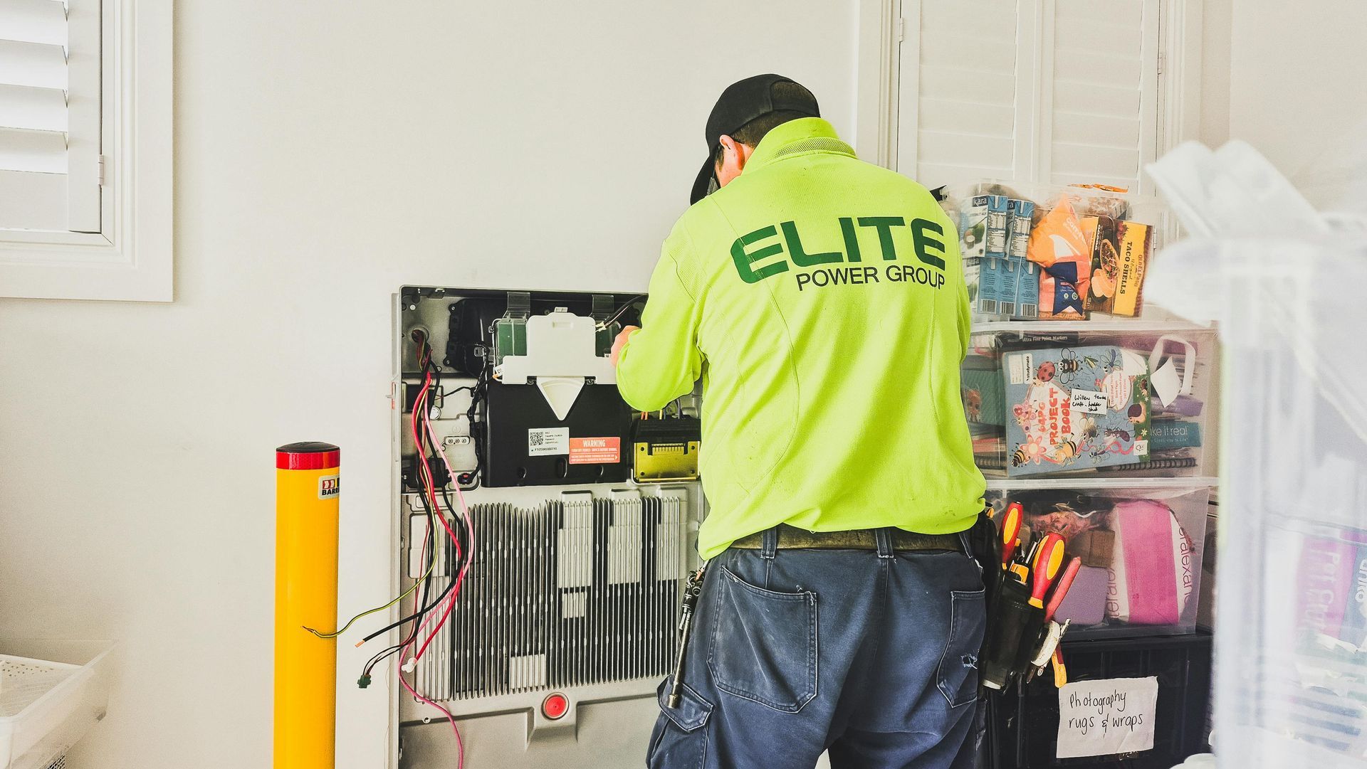 Technician in a neon green shirt works on electrical components near a yellow pole, likely installing.