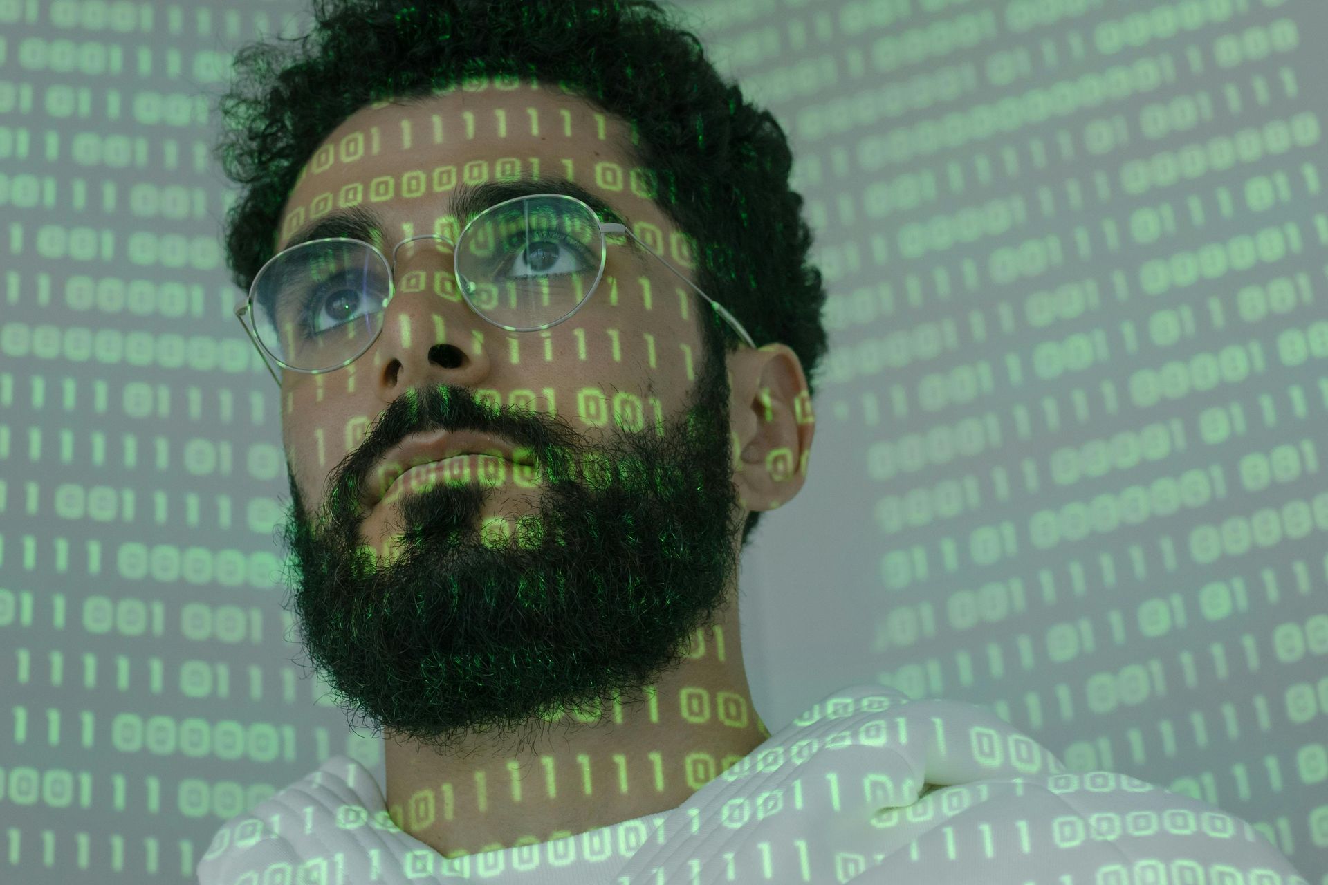 Man with glasses and beard, looking up, with binary code projected on him and the background.