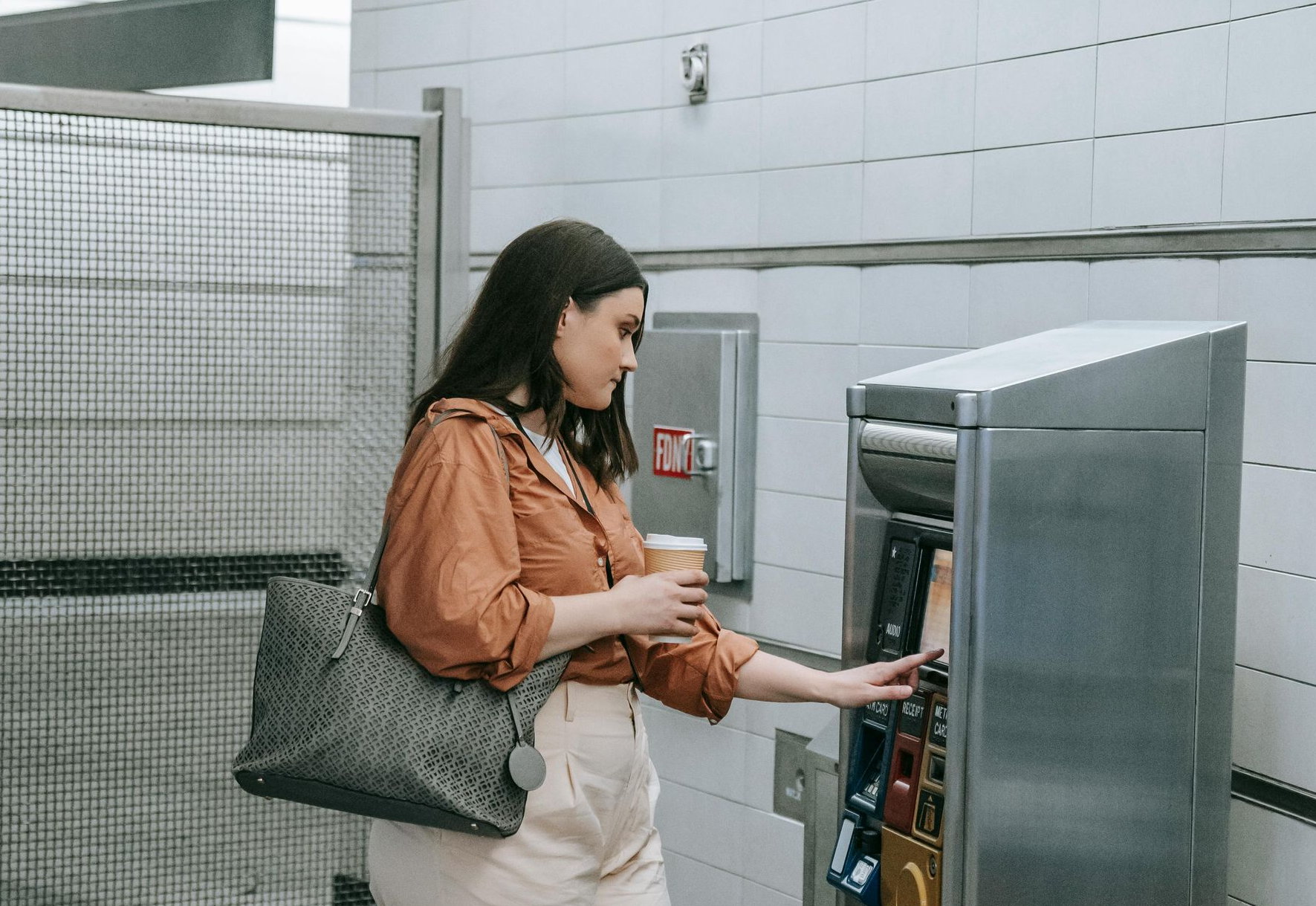 Woman at ticket machine in a subway station, holding a coffee and a bag, touching the screen.