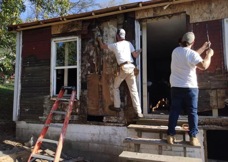 Two men working on a dilapidated red building, removing siding. One climbs, the other pulls. Ladder and steps visible.