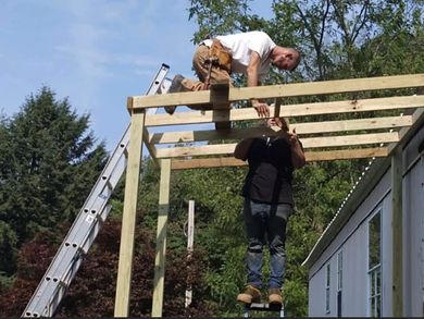 Two construction workers building a wooden structure outdoors. One on the roof, the other hanging below. Sunny day.