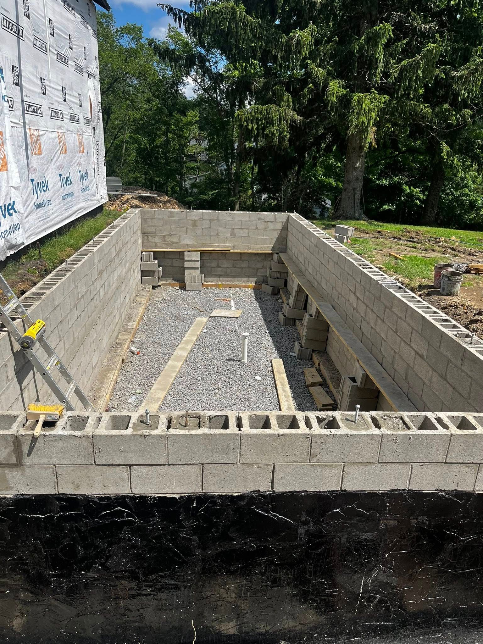 A rectangular concrete block foundation under construction with gravel base, next to a building and trees.