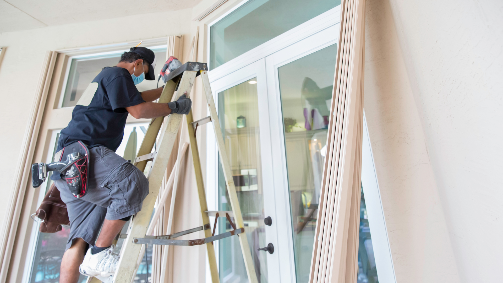 Man on ladder cleaning a window. He wears a mask, working indoors near curtains.