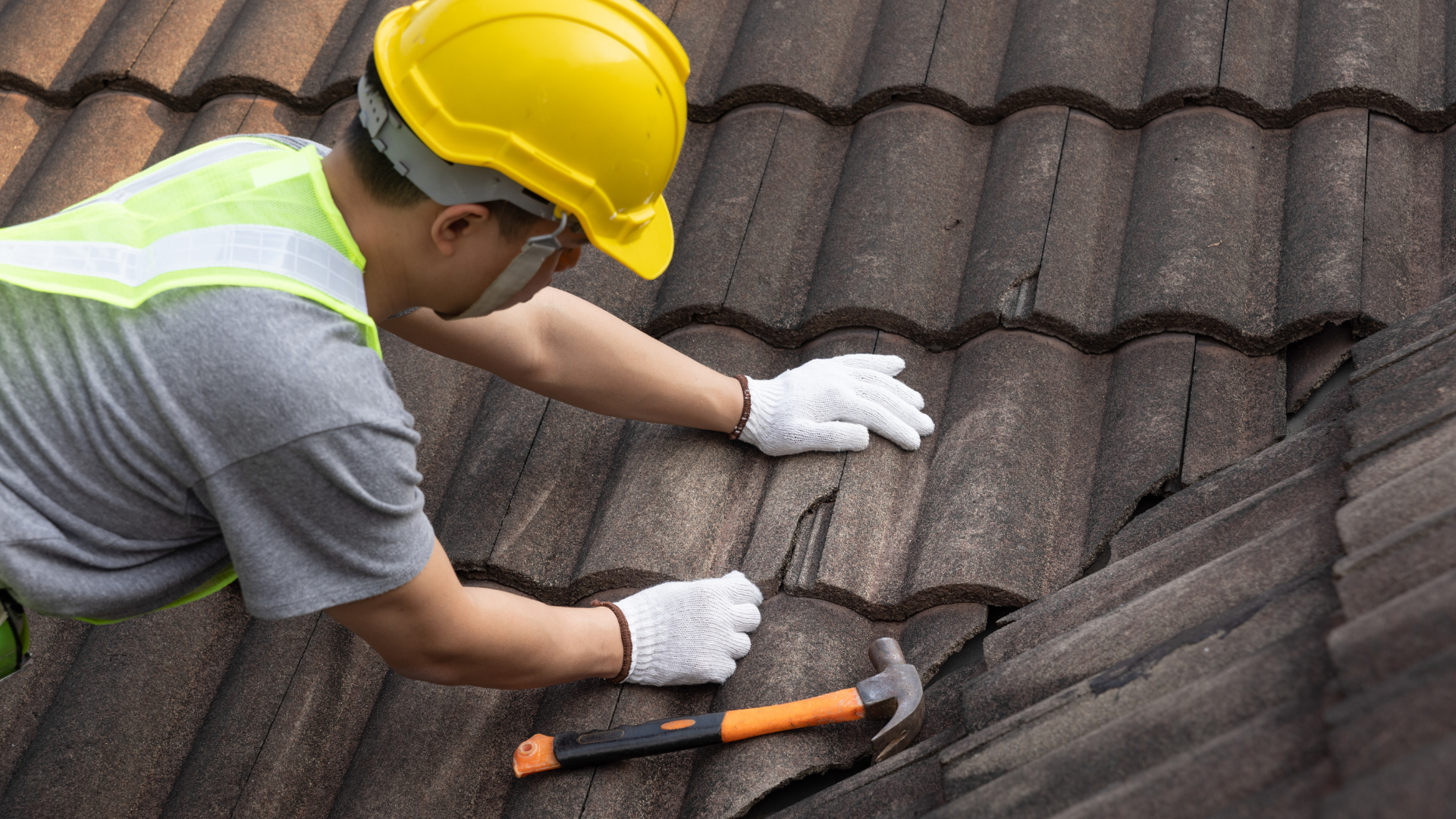 Roofer in yellow hard hat and gloves repairing damaged tile roof with a hammer.