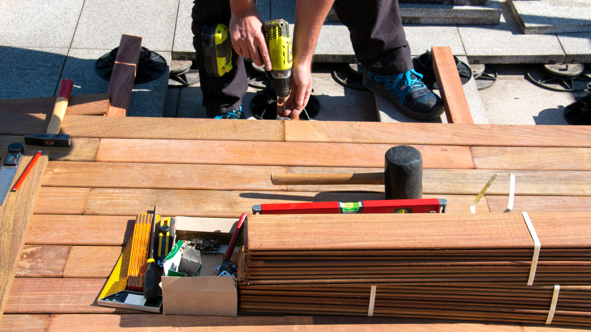 Person using a drill to install wooden deck boards, tools and materials nearby on an outdoor patio.