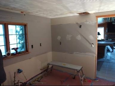 Room under renovation: gray and tan walls, exposed electrical boxes, small ladder, window with tape, doorway.