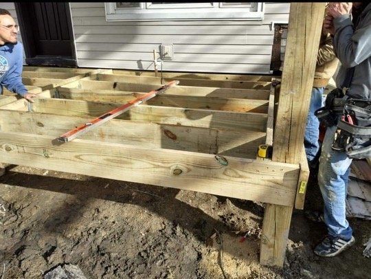 Deck frame under construction; workers in blue jeans and a house with white siding in background.
