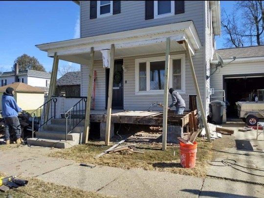 Two workers remodel a house porch; brown wood supports the roof, bare deck.