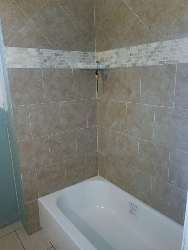 Corner shower with beige tiled walls, white tub, and decorative mosaic tile band.
