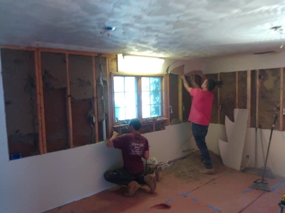 Two people installing drywall in a room with exposed studs and a window.