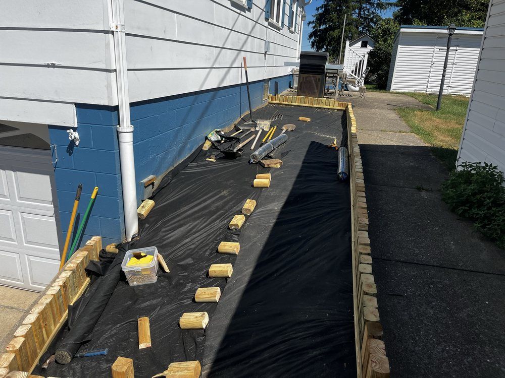 Construction site with a long, black-covered ramp bordered by wooden planks, next to a blue house and paved walkway.