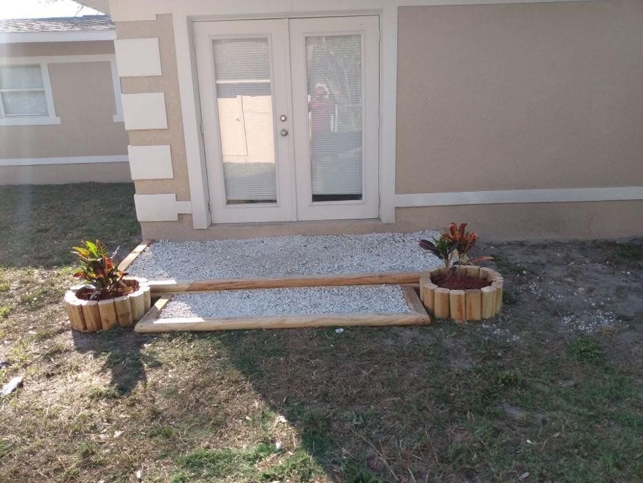 Exterior of a beige house with french doors. A gravel ramp leads to the doors, flanked by planters with plants.