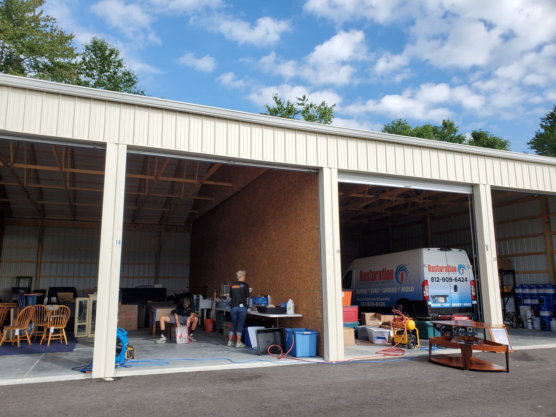 People working in an open-bay storage facility with boxes and an ice cream van parked inside under a sunny blue sky.