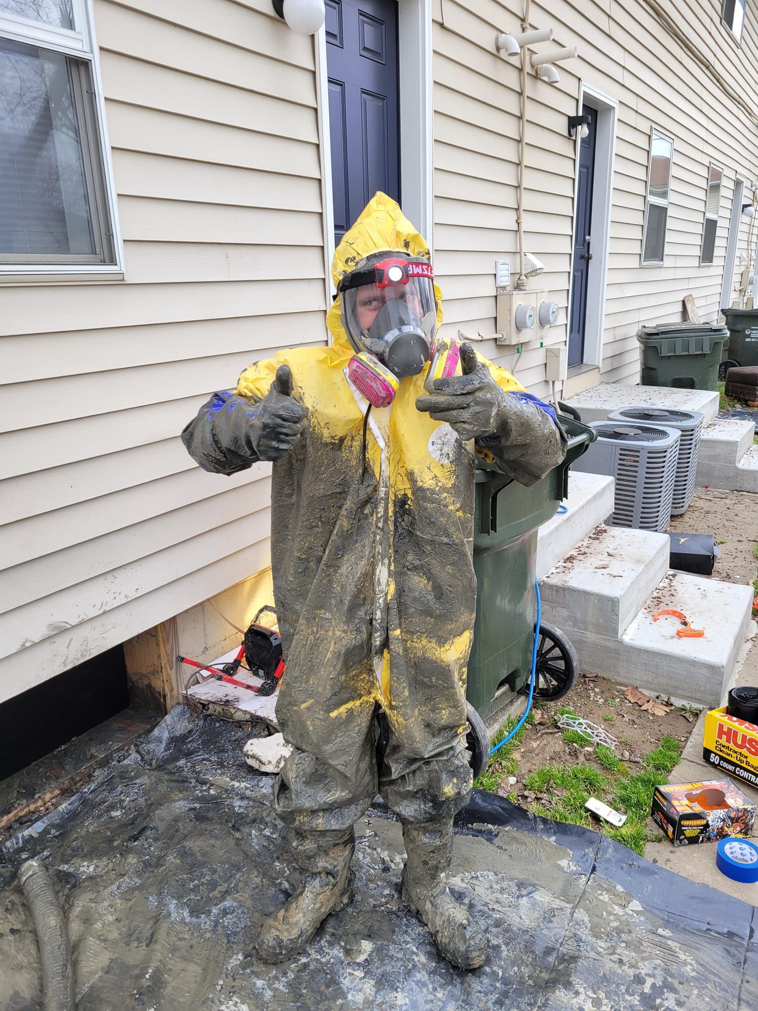 A person in a mud-covered yellow hazmat suit and respirator stands outside giving a thumbs-up.