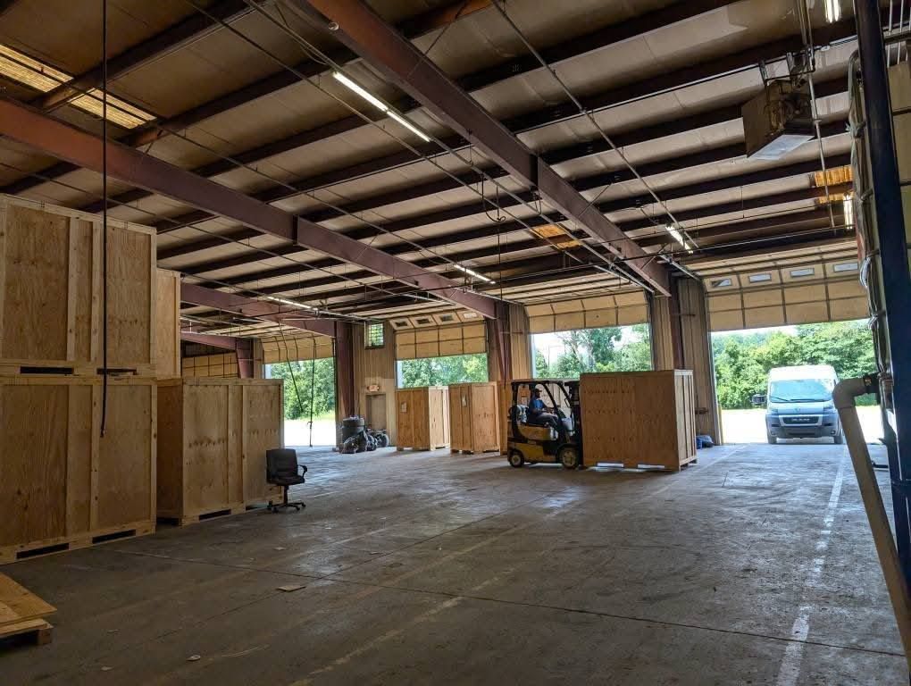 A forklift transports a wooden crate inside a spacious warehouse with open loading bays and a parked van outside.