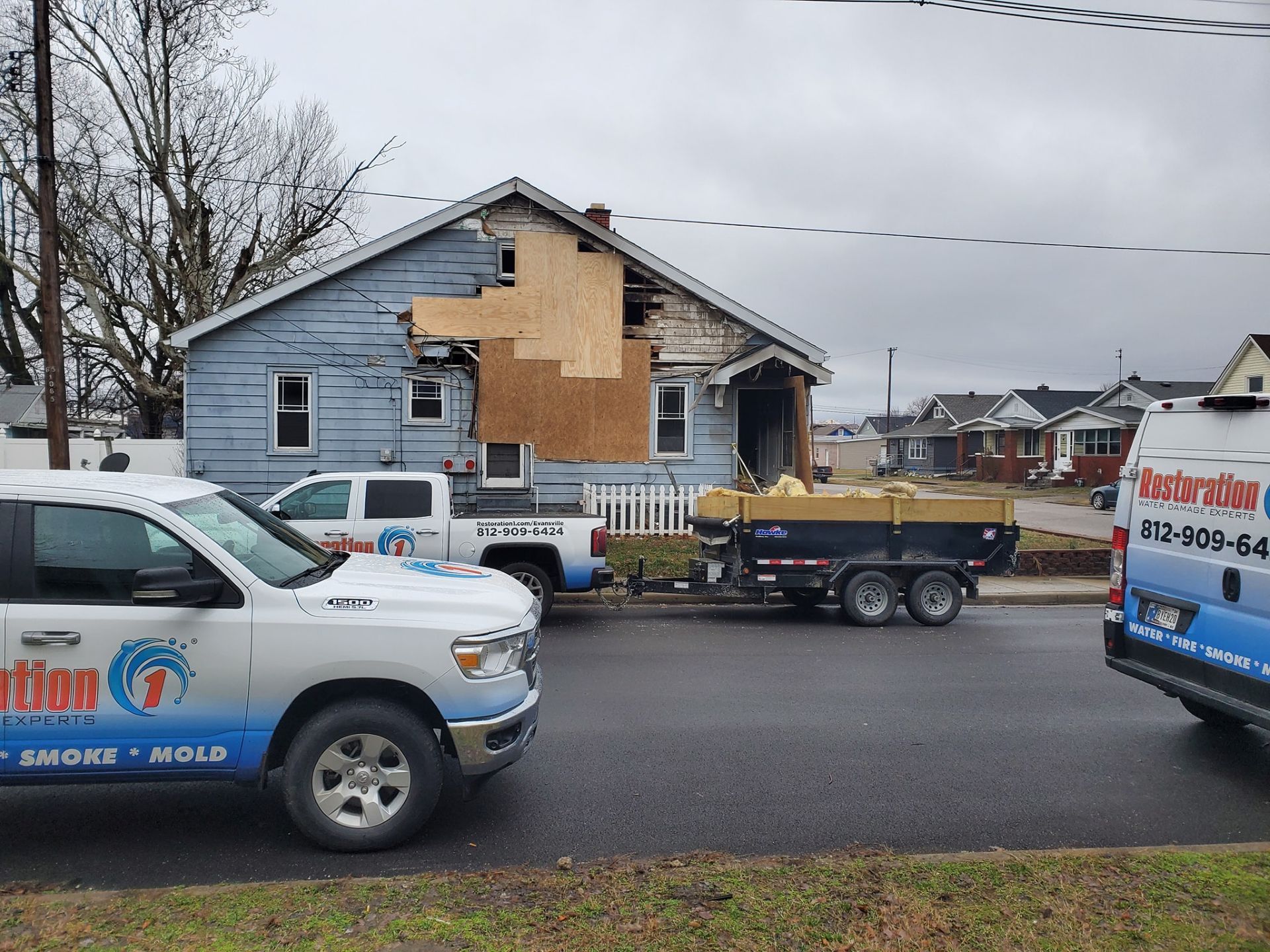 Restoration vehicles and a trailer parked on a street in front of a house with fire damage and boarded-up siding.