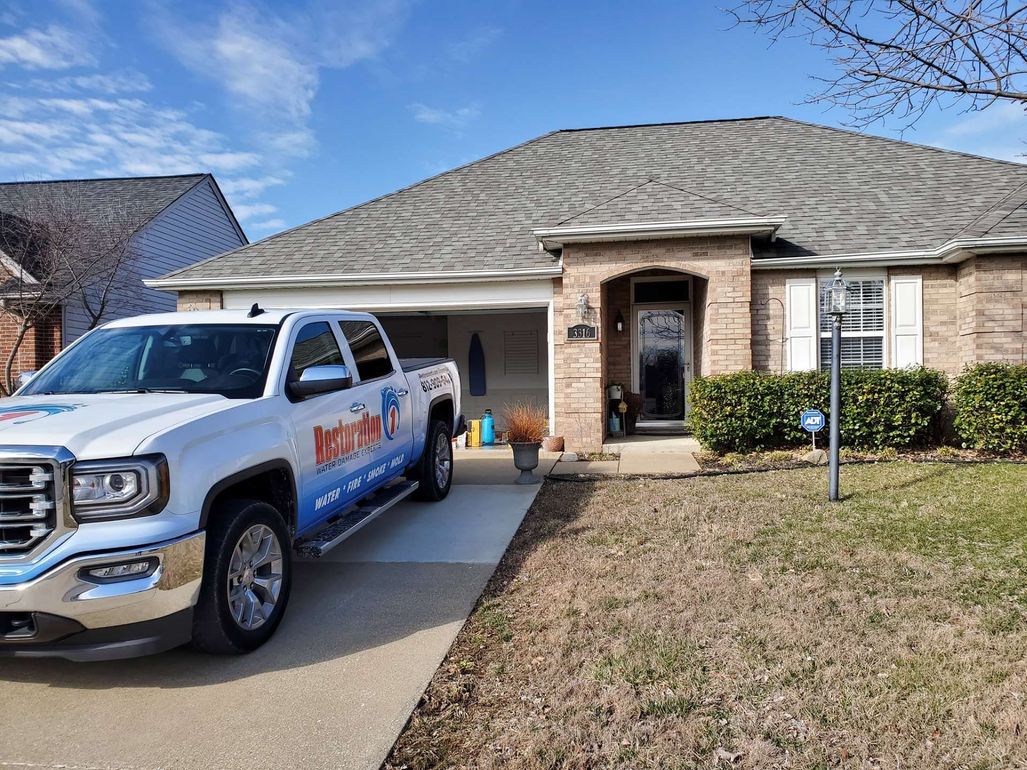 A white service truck parked in the driveway of a single-story suburban brick house under a blue, sunny sky.
