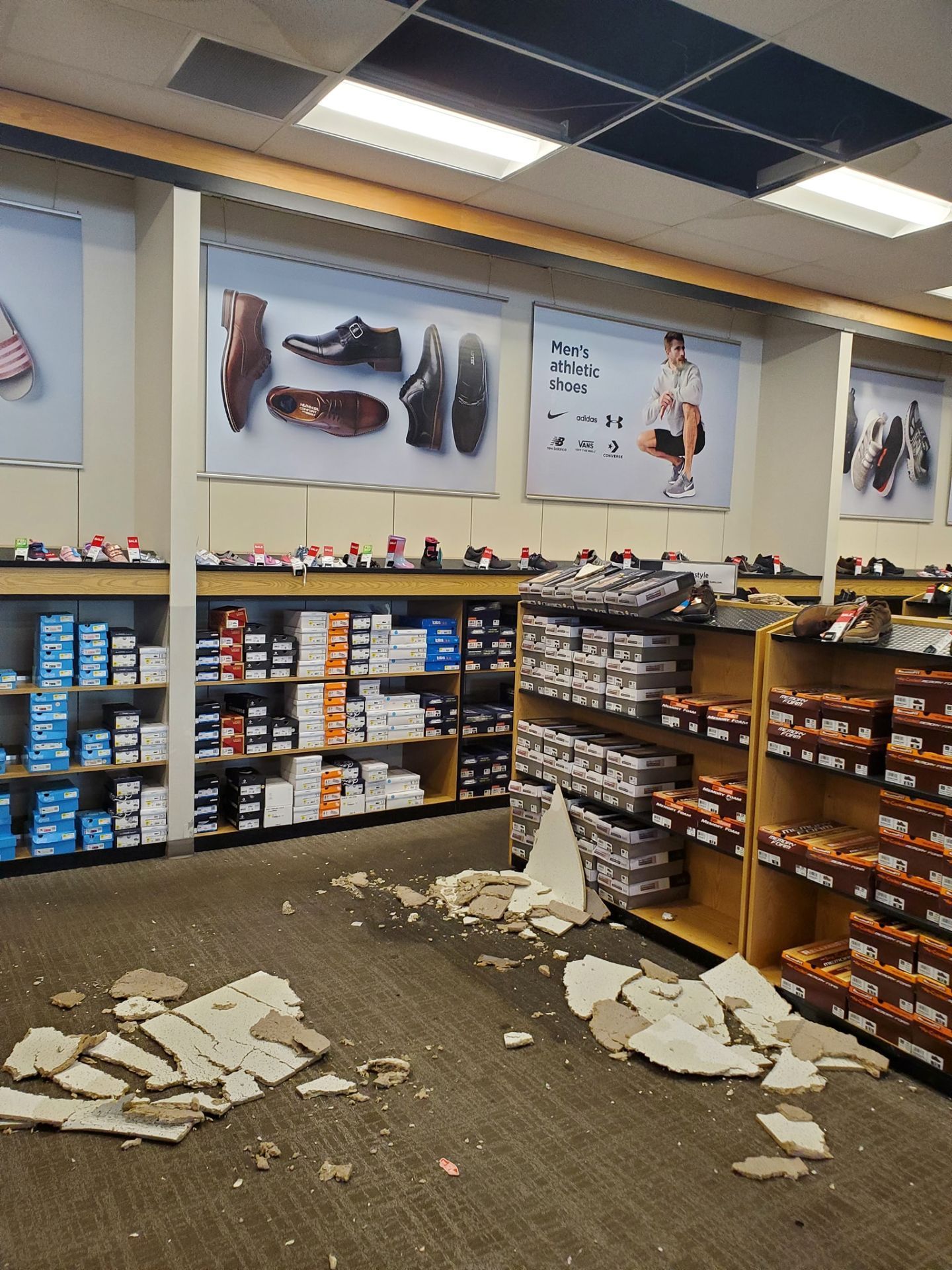 Debris from a collapsed ceiling tile lies scattered across the carpeted floor of a shoe store aisle.