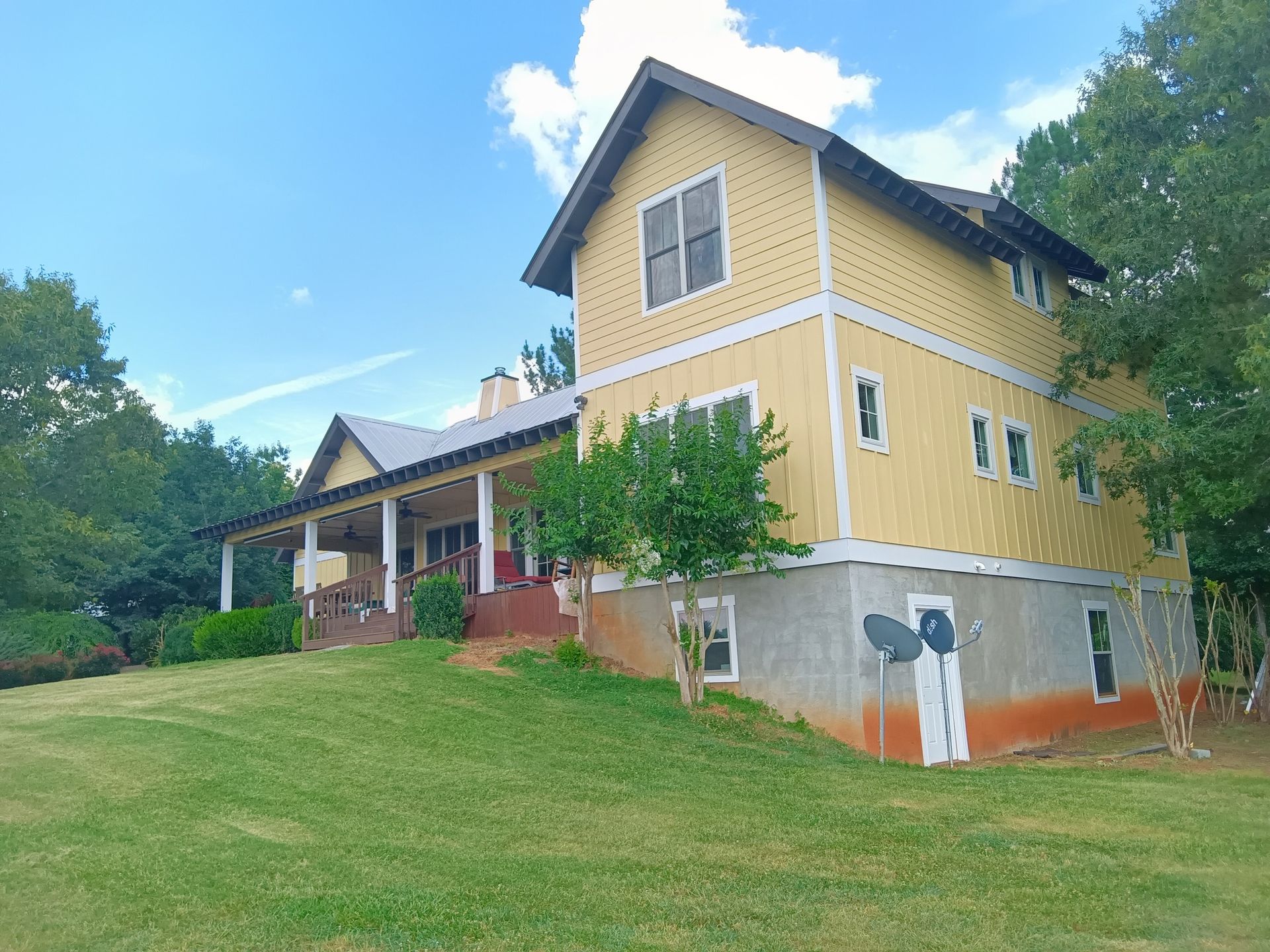 Yellow house with a wraparound porch on a grassy hill, under a blue sky.