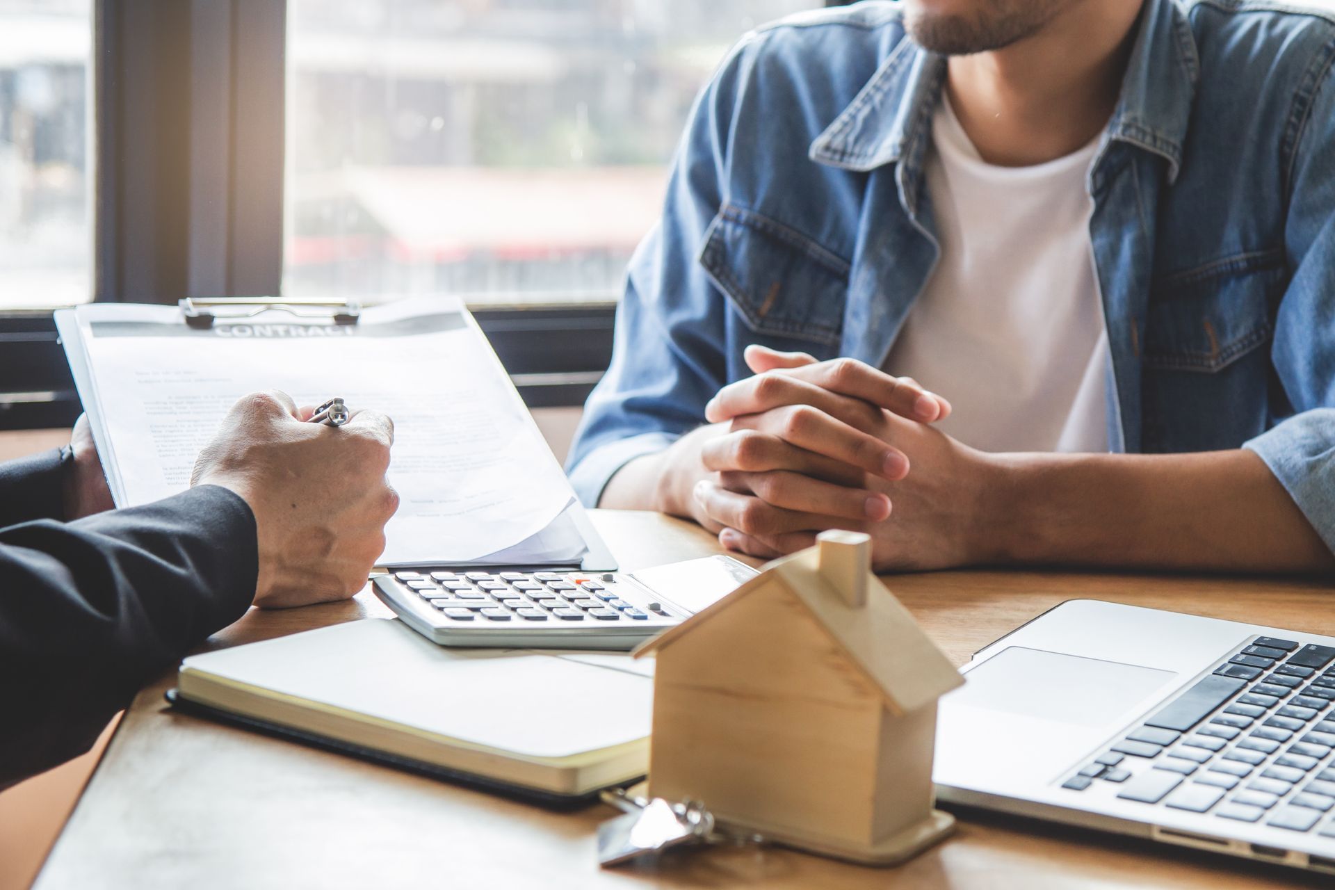 A person in a business suit reviewing documents with a client at a desk featuring a small wooden model house.