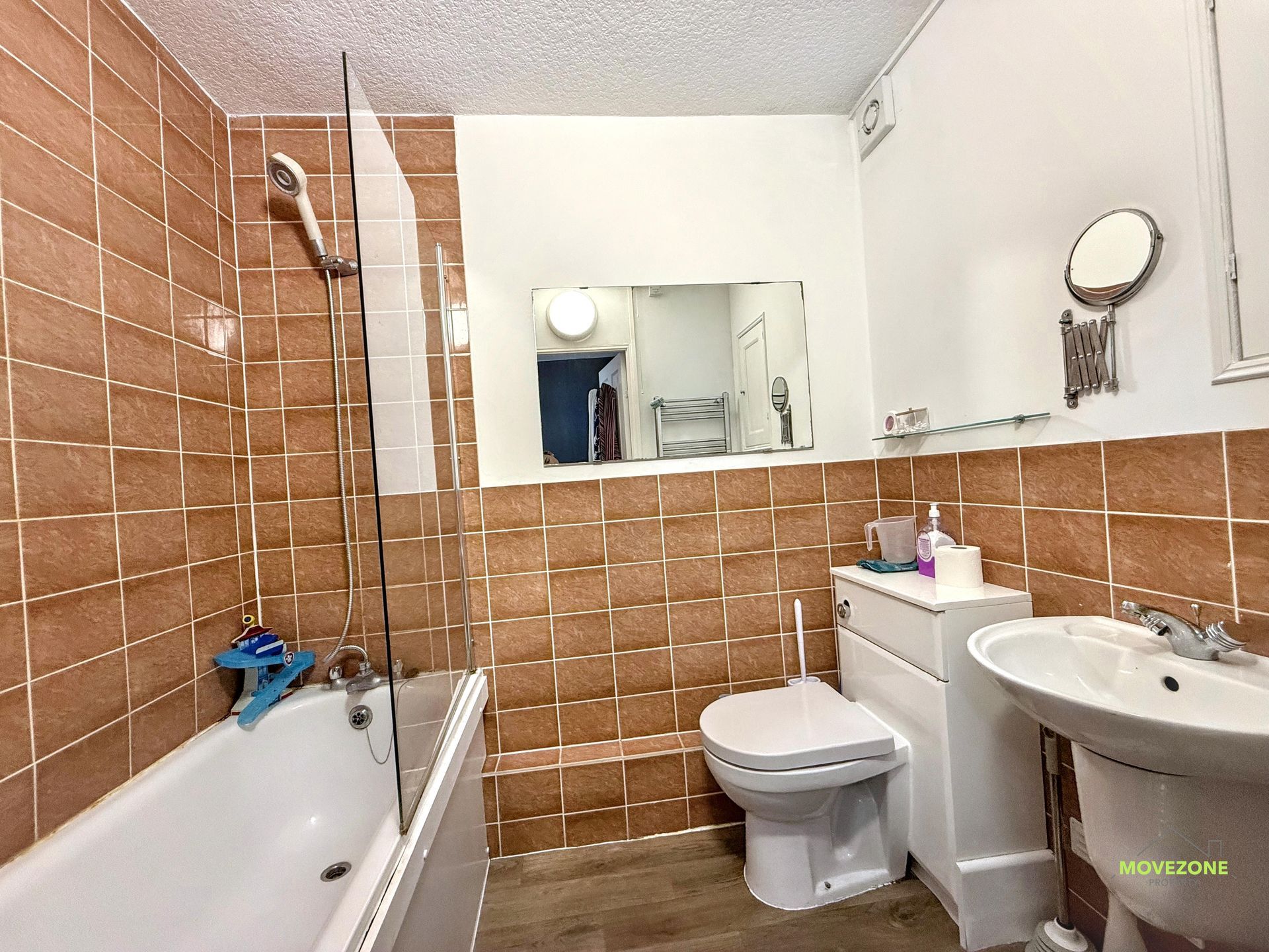 Compact bathroom with tan tiled walls, bathtub, toilet, sink, and large mirror.