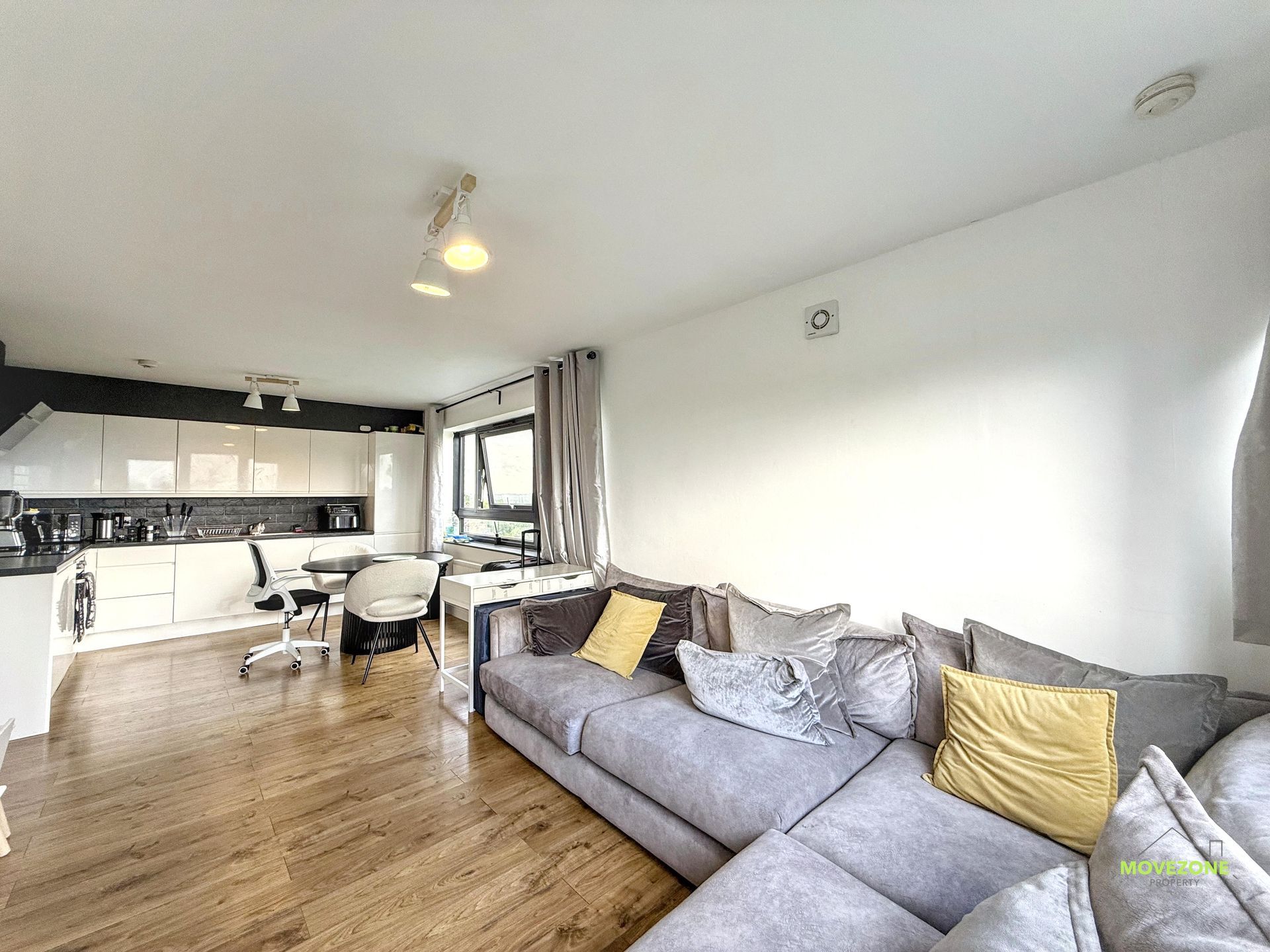 Bright open-plan living room with gray sofa, dining table, kitchen, and large windows.
