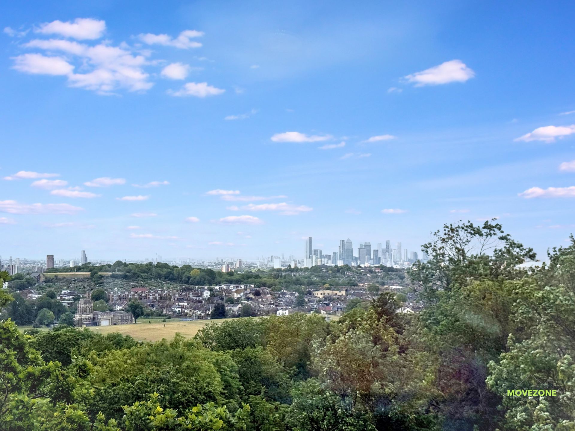 Panoramic city skyline beyond green trees and fields under a bright blue sky with scattered clouds