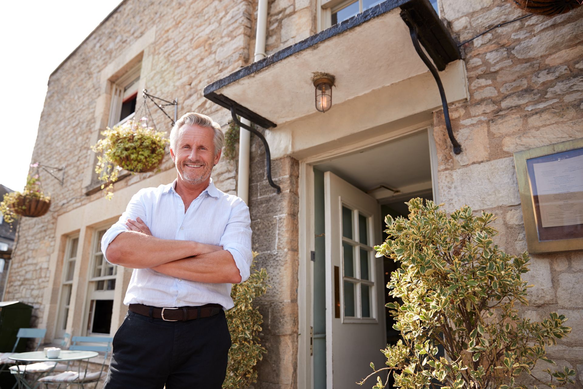 Man with arms crossed stands in front of a stone building with a doorway and awning.