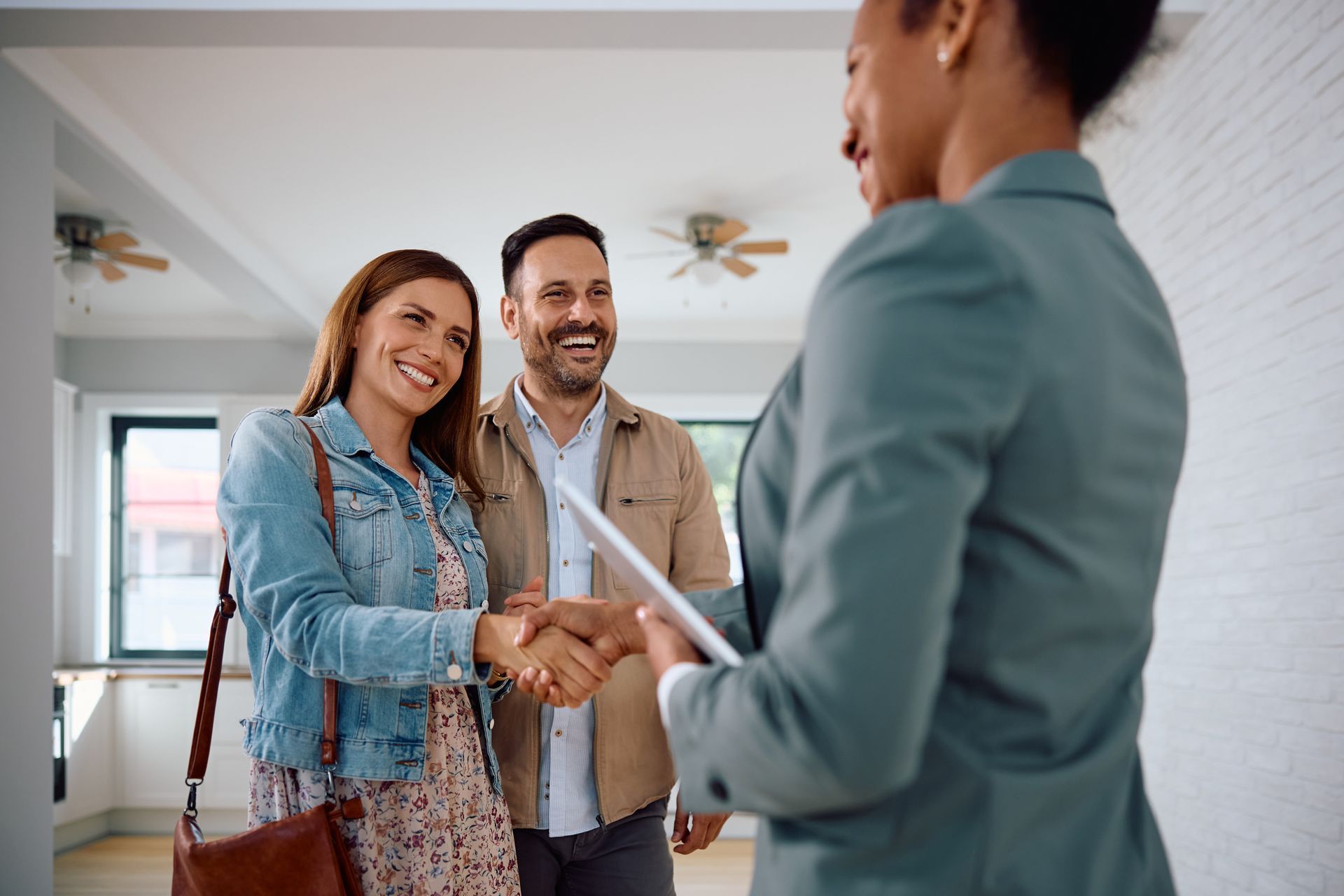 A real estate agent shakes hands with a smiling couple inside a bright, unfurnished house.